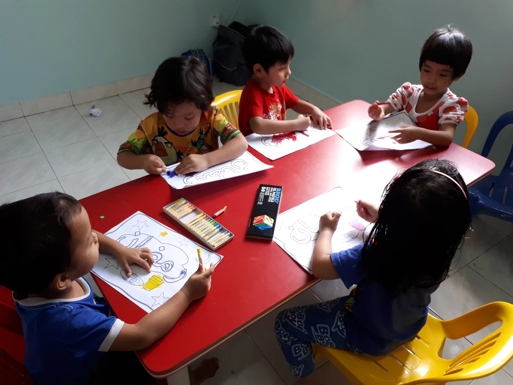 Lima kanak-kanak mewarna bersama di sekeliling meja merah.
